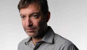 Φωτογραφία - πορτραίτο ενός άνδρα γύρω στα 45 με καστανά κοντά μαλλιά και πράσινα μάτια. Έχει κοντά αξύριστα γκριζαρισμένα μούσια και φορά γκρι πουκάμισο. Το πρόσωπό του είναι στραμμένο ελαφρά προς τα αριστερά και προς τα εκεί εστιάζει και το ήρεμο βλέμμα του.