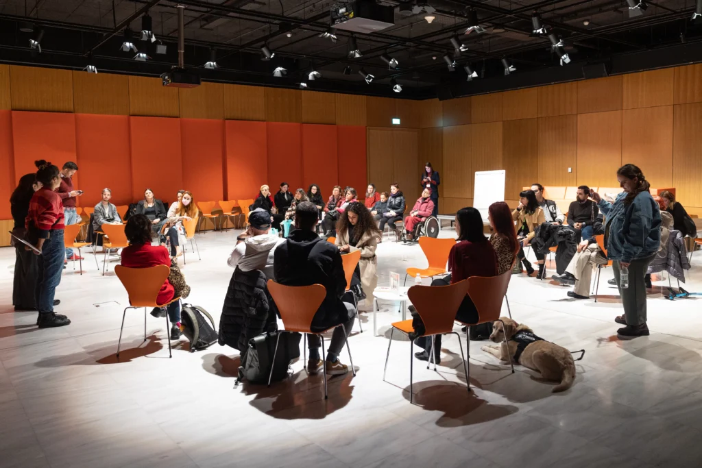 Σε μια μεγάλη αίθουσα με μαρμάρινο λευκό πάτωμα και ξύλινα πανέλα στους τοίχους, καρέκλες είναι τοποθετημένες σε τέσσερις κύκλους. Τα άτομα που παρευρίσκονται, έχουν στραμμένα τα σώματά τους προς τη μία πλευρά της αίθουσας όπου δύο όρθιες γυναίκες έχουν τον λόγο.