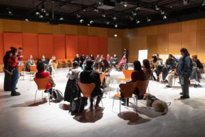 Σε μια μεγάλη αίθουσα με μαρμάρινο λευκό πάτωμα και ξύλινα πανέλα στους τοίχους, καρέκλες είναι τοποθετημένες σε τέσσερις κύκλους. Τα άτομα που παρευρίσκονται, έχουν στραμμένα τα σώματά τους προς τη μία πλευρά της αίθουσας όπου δύο όρθιες γυναίκες έχουν τον λόγο.