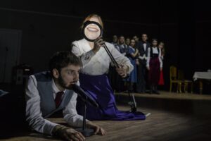 Ένας νεαρός άντρας στηρίζεται στους αγκώνες του και μιλά σε ένα μικρόφωνο στο ξύλινο πάτωμα. Δίπλα του μια γυναίκα με μπλε φούστα κρατά ένα μεγεθυντικό φακό στο στόμα της κάνοντας το χαμόγελό της να φαντάζει τεράστιο. Πίσω τους, στο βάθος της σκηνής, μια ομάδα ανθρώπων συσπειρωμένη στην άκρη τους παρακολουθεί.