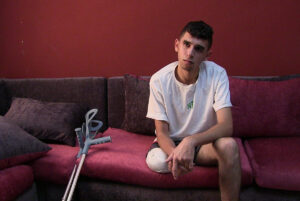 Σε έναν καφετί καναπέ μπροστά από έναν κόκκινο τοίχο, ένα νεαρό αγόρι με καστανά κοντά μαλλιά και καλοκαιρινά ρούχα κοιτά απέναντί του σα να ακούει κάποιον να του μιλά. Το ένα του πόδι, κομμένο ως το γόνατο, είναι τυλιγμένο με επιδέσμους, ενώ δίπλα του στο μαξιλάρι τού καναπέ στηρίζονται οι δυο του γκρι πατερίτσες.