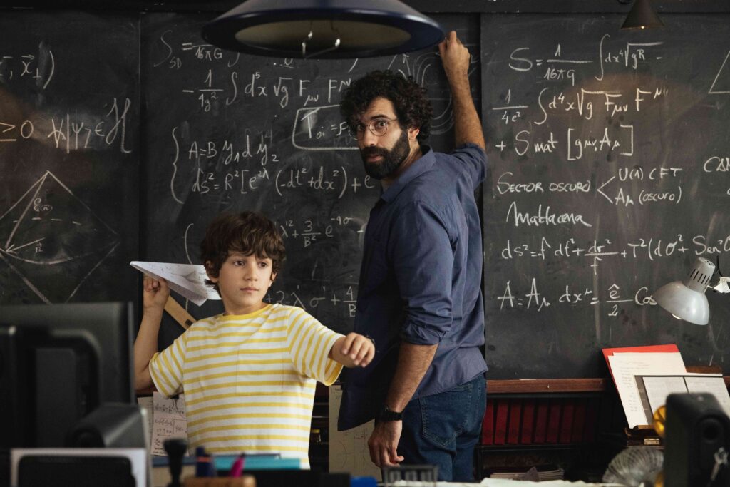 Ένας νεαρός άνδρας με σγουρά καστανά μαλλιά, γυαλιά οράσεως και μούσια γυρνά και κοιτά προς τον φακό ενώ γράφει με κιμωλία περίπλοκες μαθηματικές εξισώσεις σε έναν μαυροπίνακα. Ένα αγόρι γύρω στα 10 μπροστά του ετοιμάζεται να πετάξει μια χάρτινη σαΐτα.