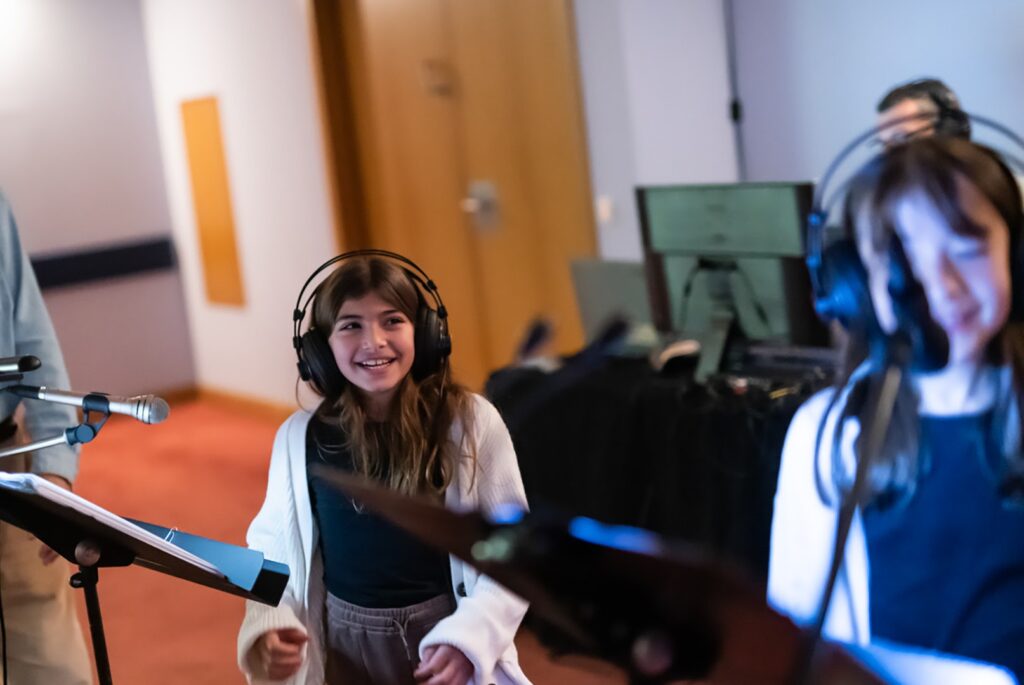 Δύο κορίτσια γύρω στα 12 φορούν ακουστικά και μιλούν γελώντας μπροστά σε μικρόφωνα και αναλόγια στημένα μπροστά τους.