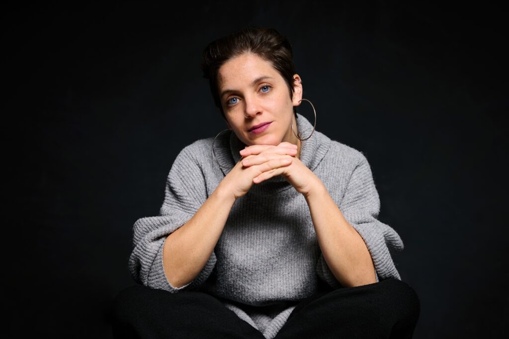Σε μαύρο φόντο και σε πλάνο από τη μέση και πάνω, η Νάγια, μία λευκή γυναίκα με μπλε μάτια και κοντά μαύρα μαλλιά, κοιτάει την κάμερα με ήρεμο ύφος. Φοράει ένα γκρι πουλόβερ και ένα μεγάλο κρίκο στο αριστερό αυτί. Έχει δεμένες τις παλάμες μεταξύ τους, κάτω από το πηγούνι της.