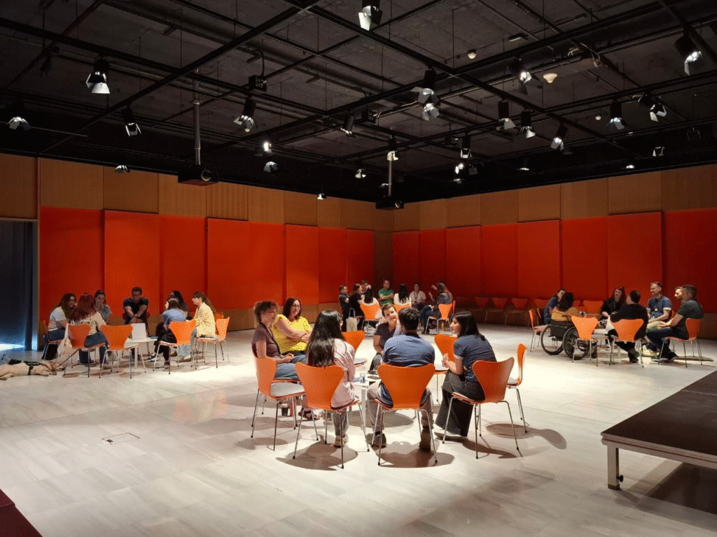 Σε μια σύγχρονη, ευρύχωρη αίθουσα με ψηλό ταβάνι, ομάδες ανθρώπων κάθονται γύρω από στρογγυλά τραπέζια κάτω από κρεμαστά φωτιστικά. Οι έντονες πορτοκαλί καρέκλες τους εναρμονίζονται με τα μεγάλα πορτοκαλί πάνελ στους τοίχους, δημιουργώντας ένα ζωντανό φόντο. Οι συμμετέχοντες φαίνεται να είναι πλήρως αφοσιωμένοι στις συζητήσεις τους.