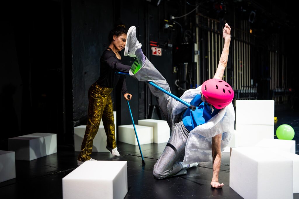 Σε θεατρική σκηνή με διάσπαρτους λευκούς κύβους, δύο γυναίκες εκτελούν μια σύνθετη χορευτική κίνηση. Η γυναίκα στο μπροστινό μέρος, με ροζ κράνος και ασημί κοστούμι, στηρίζει το σώμα της στο αριστερό της χέρι και γόνατο τεντώνοντας τα υπόλοιπα μέλη ψηλά. Το δεξί της πόδι ακουμπά την υψωμένη μπλε πατερίτσα της άλλης γυναίκας που στέκεται όρθια μπροστά της με χρυσό παντελόνι και μαύρη μπλούζα.