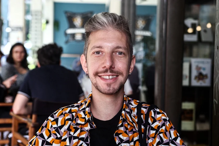 Ένας νεαρός άνδρας με γαλάζια μάτια και μια γκρίζα τούφα στο κέντρο της φράντζας των κοντών μαλλιών του χαμογελά απαλά κοιτώντας τον φακό. Φορά εμπριμέ πουκάμισο και μαύρη μπλούζα από μέσα, έχει τρύπες στα αυτιά και στη μύτη και κάθεται σε χώρο που θυμίζει καφέ.