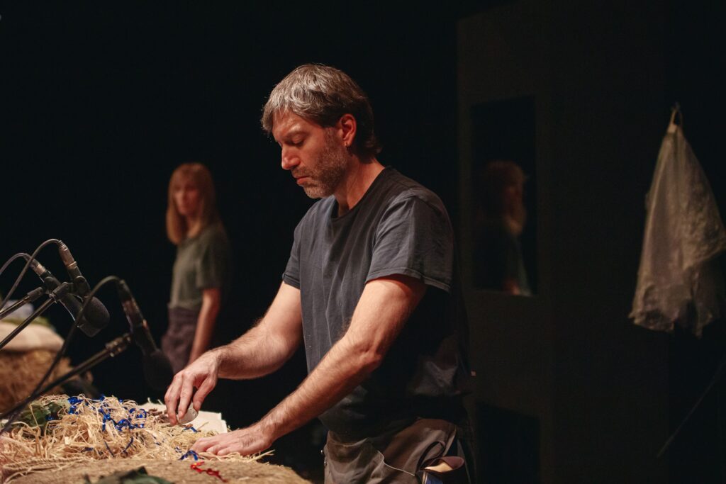 Ένας άνδρας γύρω στα 45 με μαύρο t-shirt ψηλαφίζει ξερά χόρτα και μερικές μπλε πλαστικές γιρλάντες που βρίσκονται σε κουβάρι μπροστά του κάτω από μικρόφωνα. Στο βάθος, μια ξανθιά γυναίκα, εξίσου απλά ντυμένη, παρακολουθεί τις κινήσεις του. Ο χώρος γύρω τους είναι σκοτεινός, το φως είναι εστιασμένο μόνο πάνω στον άνδρα.