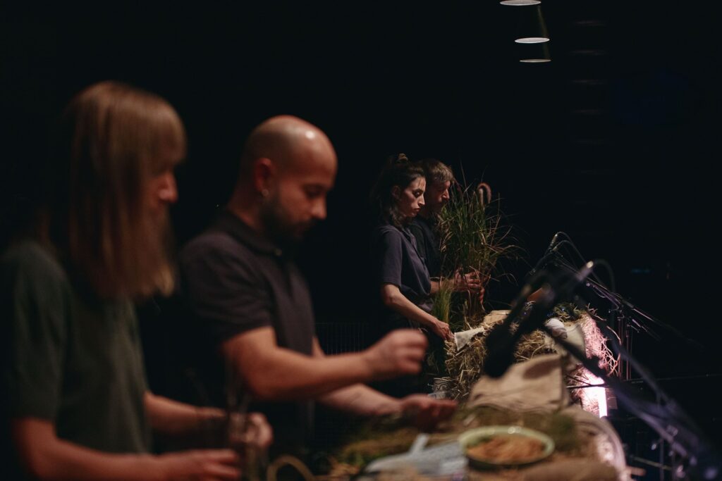 Σε έναν σκοτεινό σκηνικό χώρο, τέσσερα άτομα ντυμένα με σκούρα ρούχα στέκονται στη σειρά πάνω από δεμάτια στάχυα και άλλα μικροαντικείμενα. Μπροστά τους μικρόφωνα είναι στραμμένα στο ύψος των χεριών τους καθώς αυτά κινούνται και διαδρούν με τα αντικείμενα.