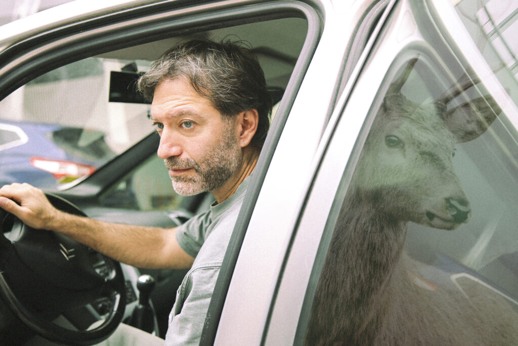 Ένας γκριζομάλλης άνδρας οδηγά με ανοιχτό παράθυρο ένα αυτοκίνητο. Στην αντανάκλαση του πίσω τζαμιού φαίνεται το κεφάλι ενός ελαφιού. Εκείνος και το ελάφι μοιάζουν να συνδέονται με κάποιο μυστήριο τρόπο καθώς τα κεφάλια τους σχεδόν εφάπτονται μέσα από αυτήν την οπτική ψευδαίσθηση που δημιουργείται.