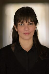 Μια νεαρή γυναίκα με αφέλειες, μακριά καστανά μαλλιά και καστανά μάτια ποζάρει κοιτώντας ευθεία τον φακό. Ένα αμυδρό χαμόγελο απλώνεται στα χείλη της. Φορά μαύρο πουκάμισο και μαύρο ζακετάκι.