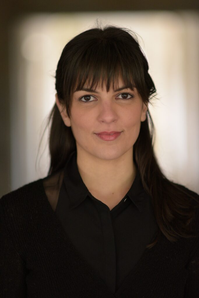 Μια νεαρή γυναίκα με αφέλειες, μακριά καστανά μαλλιά και καστανά μάτια ποζάρει κοιτώντας ευθεία τον φακό. Ένα αμυδρό χαμόγελο απλώνεται στα χείλη της. Φορά μαύρο πουκάμισο και μαύρο ζακετάκι.