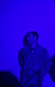 Ένας άνδρας με πουκάμισο στέκεται μπροστά σε τοίχο κάτω από έντονα μπλε φώτα λες και βρίσκεται σε συναυλία. Το βλέμμα του εστιάζει κάπου δεξιά και χαμογελά σα να παρακολουθεί κάτι που τον έχει συνεπάρει.