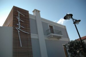 Στην εξωτερική επιφάνεια του κτιρίου που στεγάζει το Μουσείο Καζαντζάκη, πάνω σε κάθετο στόρυ από λεπτά οριζόντια ξύλινα δοκάρια, εμφανίζεται τρισδιάστατη και τεράστια η χειρόγραφη υπογραφή του συγγραφέα με τα καλλιγραφικά του γράμματα τυπωμένα κάθετα σε λευκό χρώμα.