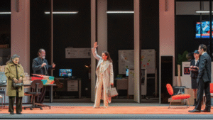 Σε έναν μεγάλο χώρο εργασίας με γραφεία, καρέκλες, οθόνες και ντοσιέ γύρω γύρω, μια γυναίκα με κρεμ καπαρντίνα χαιρετά με το χέρι ψηλά τους παρευρισκόμενους σαν μόλις να μπήκε στον χώρο. Τα υπόλοιπα πρόσωπα χαμογελούν, εκτός από μια μεγαλύτερης ηλικίας γυναίκα στο μπροστινό μέρος που στέκει σκυθρωπή με την τσάντα της στα χέρια.