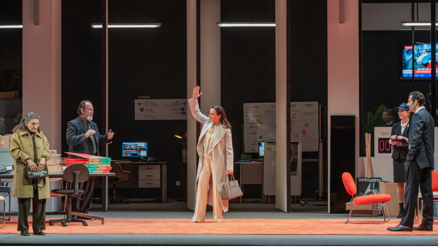 Σε έναν μεγάλο χώρο εργασίας με γραφεία, καρέκλες, οθόνες και ντοσιέ γύρω γύρω, μια γυναίκα με κρεμ καπαρντίνα χαιρετά με το χέρι ψηλά τους παρευρισκόμενους σαν μόλις να μπήκε στον χώρο. Τα υπόλοιπα πρόσωπα χαμογελούν, εκτός από μια μεγαλύτερης ηλικίας γυναίκα στο μπροστινό μέρος που στέκει σκυθρωπή με την τσάντα της στα χέρια.