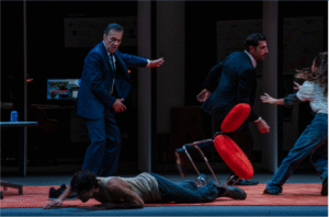 Σε έναν χώρο εργασίας επικρατεί ταραχή. Ένας νεαρός άνδρας με φανελάκι είναι πεσμένος μπρούμυτα στο πάτωμα. Από πάνω του, ένας κοστουμαρισμένος άνδρας τον κοιτά αιφνιδιασμένος καθώς ένας τρίτος άνδρας με κοστούμι στα δεξιά φεύγει με φόρα παρασύροντας στον διάβα του μια κόκκινη καρέκλα που είναι έτοιμη να πέσει. Μια νεαρή κοπέλα με τζιν μοιάζει να προσπαθεί να τον σταματήσει με τα χέρια της τεντωμένα προς το μέρος του.