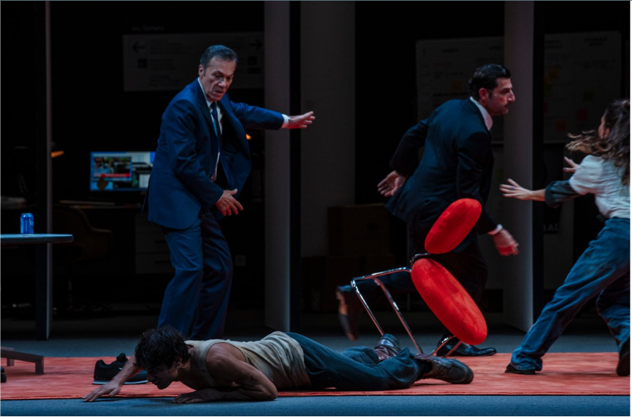 Σε έναν χώρο εργασίας επικρατεί ταραχή. Ένας νεαρός άνδρας με φανελάκι είναι πεσμένος μπρούμυτα στο πάτωμα. Από πάνω του, ένας κοστουμαρισμένος άνδρας τον κοιτά αιφνιδιασμένος καθώς ένας τρίτος άνδρας με κοστούμι στα δεξιά φεύγει με φόρα παρασύροντας στον διάβα του μια κόκκινη καρέκλα που είναι έτοιμη να πέσει. Μια νεαρή κοπέλα με τζιν μοιάζει να προσπαθεί να τον σταματήσει με τα χέρια της τεντωμένα προς το μέρος του.