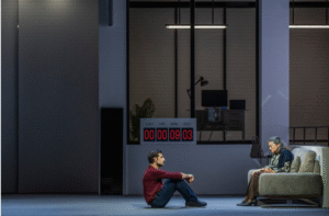 Μια γυναίκα γύρω στα 80 με γυαλιά και ένα ζακετάκι ριγμένο στους ώμους της. κάθεται σε έναν γκρίζο καναπέ τοποθετημένο κάθετα στον χώρο. Μιλά μπροστά της σε έναν νεαρότερο άνδρα με μπορντό μπλούζα και μπλε παντελόνι που την παρακολουθεί προσεκτικά καθισμένος στο πάτωμα. Καθώς οι δυο τους συνομιλούν σε προφίλ, πίσω τους στο βάθος, ένα χρονόμετρο με κόκκινα ψηφία δείχνει 9 λεπτά και 3 δευτερόλεπτα.