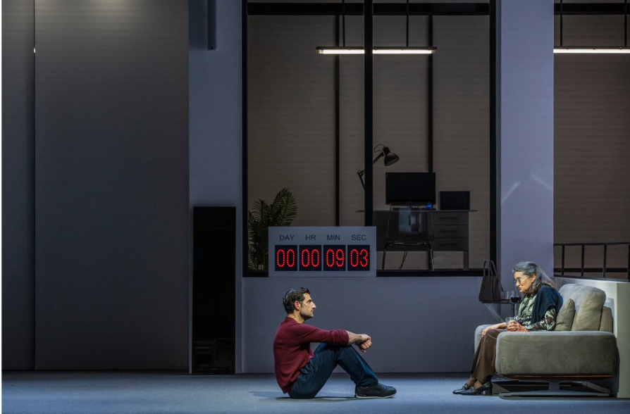 Μια γυναίκα γύρω στα 80 με γυαλιά και ένα ζακετάκι ριγμένο στους ώμους της. κάθεται σε έναν γκρίζο καναπέ τοποθετημένο κάθετα στον χώρο. Μιλά μπροστά της σε έναν νεαρότερο άνδρα με μπορντό μπλούζα και μπλε παντελόνι που την παρακολουθεί προσεκτικά καθισμένος στο πάτωμα. Καθώς οι δυο τους συνομιλούν σε προφίλ, πίσω τους στο βάθος, ένα χρονόμετρο με κόκκινα ψηφία δείχνει 9 λεπτά και 3 δευτερόλεπτα. 