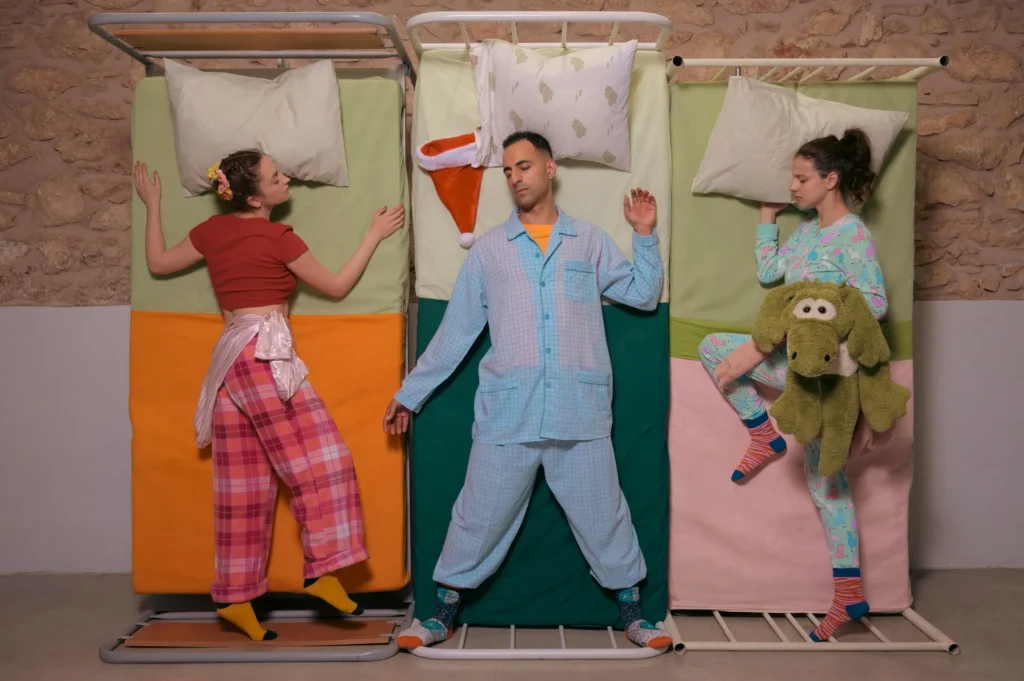 Φωτογραφία τραβηγμένη από ψηλά όπου τρία νεαρά άτομα με πιτζάμες είναι ξαπλωμένα με κλειστά μάτια σε τρία κρεβάτια στη σειρά. Δυο νεαρές γυναίκες κοιμούνται στα ακριανά κρεβάτια κι ένας νεαρός άνδρας στο μεσαίο. Ένα σκουφί Άγιου Βασίλη βρίσκεται δίπλα στο μαξιλάρι του. Στα δεξιά η γυναίκα αγκαλιάζει ένα μεγάλο λούτρινο αρκουδάκι και φοράει ριγέ κάλτσες και πιτζάμες, ενώ η γυναίκα στα αριστερά φοράει καρό πιτζάμες, καφέ μπλούζα και κίτρινες κάλτσες.