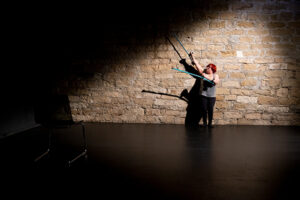 Σε έναν χώρο με πέτρινους τοίχους και μαύρο λείο πάτωμα, μια γυναίκα με κόκκινα μαλλιά στέκεται όρθια με την πλάτη της να ακουμπά τον πίσω τοίχο. Φορά γκρι φανελάκι και μαύρο κολάν και υψώνει τις δύο πατερίτσες της στον αέρα προς τα δεξιά της σα να εκτελεί κάποια χορογραφία.