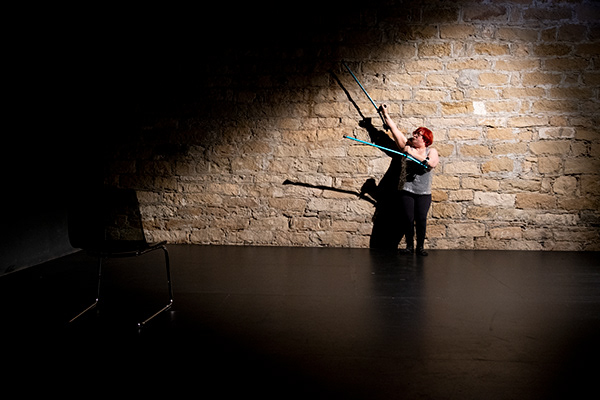 Σε έναν χώρο με πέτρινους τοίχους και μαύρο λείο πάτωμα, μια γυναίκα με κόκκινα μαλλιά στέκεται όρθια με την πλάτη της να ακουμπά τον πίσω τοίχο. Φορά γκρι φανελάκι και μαύρο κολάν και υψώνει τις δύο πατερίτσες της στον αέρα προς τα δεξιά της σα να εκτελεί κάποια χορογραφία.