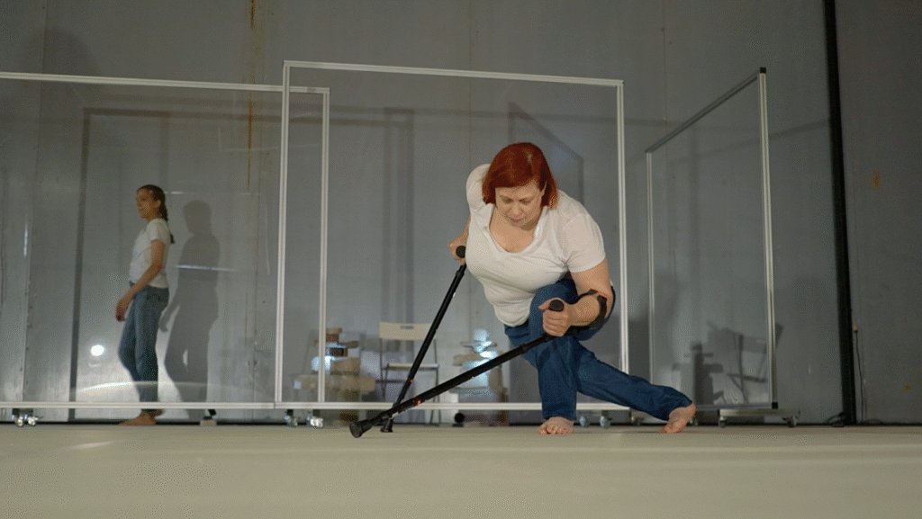 Δύο πλαίσια από τζάμι με ροδάκια στο κάτω μέρος τους χωρίζουν δύο γυναίκες σε έναν χώρο με γκρίζο πάτωμα. Οι δύο γυναίκες φορούν μπλε τζιν και λευκό t-shirt. Η μία βηματίζει στο πίσω μέρος, ενώ η άλλη μπροστά γέρνει το σώμα της στο πλάι και στηρίζεται στις πατερίτσες της διατηρώντας μια δύσκολη ισορροπία.