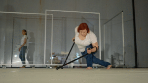 Δύο πλαίσια από τζάμι με ροδάκια στο κάτω μέρος τους χωρίζουν δύο γυναίκες σε έναν χώρο με γκρίζο πάτωμα. Οι δύο γυναίκες φορούν μπλε τζιν και λευκό t-shirt. Η μία βηματίζει στο πίσω μέρος, ενώ η άλλη μπροστά γέρνει το σώμα της στο πλάι και στηρίζεται στις πατερίτσες της διατηρώντας μια δύσκολη ισορροπία.