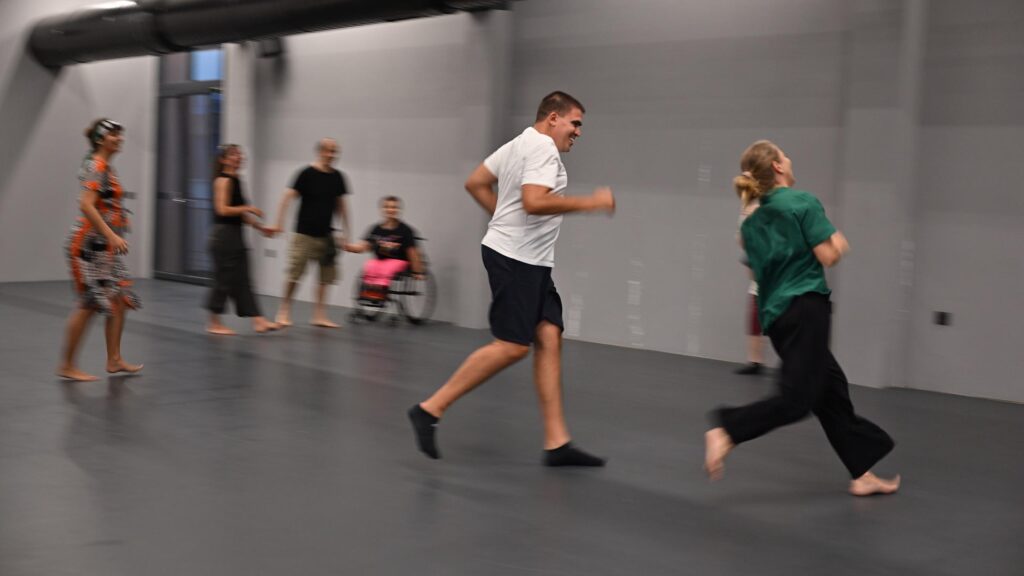 Σε μια απλοχωρη αίθουσα με γκρι πάτωμα ένας ψηλός άνδρας κυνηγά γελώντας μια ξανθιά γυναίκα. Πίσω τους στο βαθος, μια γυναίκα σε αμαξίδιο, ένας άνδρας και μια γυναίκα κρατιούνται χέρι-χέρι, ενώ μια ακόμη γυναίκα τούς παρακολουθεί με προσοχή έχοντας στρέψει το σώμα της προς αυτούς.