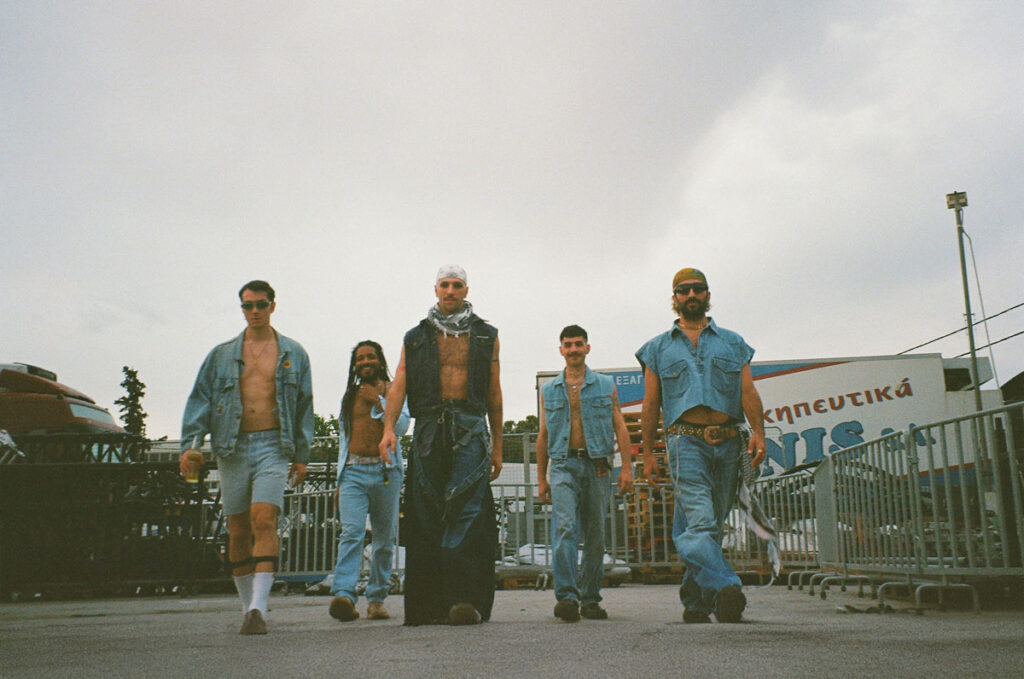 Σε εξωτερικό χώρο με κάγκελα, πέντε αγόρια με τζιν παντελόνια και τζάκετ προχωρούν προς την κάμερα με βλέμματα κεφάτα και γεμάτα αυτοπεποίθηση.