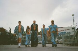 Σε εξωτερικό χώρο με κάγκελα, πέντε αγόρια με τζιν παντελόνια και τζάκετ προχωρούν προς την κάμερα με βλέμματα κεφάτα και γεμάτα αυτοπεποίθηση.