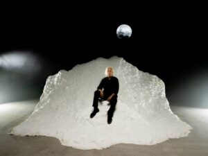 Στο σκοτάδι, ένας τεράστιος λευκός όγκος σα μια γιγάντια χιονόμπαλα ακτινοβολεί ολόλευκη σε σκηνικό χώρο. Πάνω της βρίσκεται καθισμένος ένας άνδρας με γκρίζα μαλλιά και μαύρα ρούχα. Η φιγούρα του μοιάζει μικροσκοπική πάνω στον τεράστιο λευκό όγκο. Από πάνω, ένα λευκό στρογγυλό φως, θυμίζει φεγγάρι σε συννεφιασμένο ουρανό.