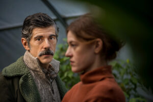 Σε ενα θερμοκήπιο με πρασινάδες, μια κοκκινομάλλα γυναίκα με πορτοκάλι πουλόβερ στέκεται προφίλ και κοιτάζει σοβαρή ευθεία μπροστά της. Πίσω της, ένας άνδρας με γκρίζα μαλλιά και μουστάκι κοιτά τον φακό σαν να μας επιτρέπει να γίνουμε μέρος μιας ιδιωτικής στιγμής.