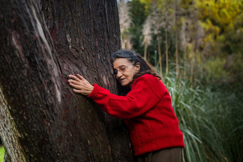 Σε ένα κατάφυτο τοπίο, μια μεγάλης ηλικίας γυναίκα με γυαλιά οράσεως και κόκκινο πουλόβερ έχει γείρει τρυφερά το σώμα της πάνω σε έναν παχύ κορμό δέντρου. Τυλίγει τα χέρια της γύρω του σε μια αγκαλιά που δεν κλείνει χαμογελώντας με κλειστά τα μάτια σα να αφήνεται ανακουφισμένη γαλήνια πάνω του.