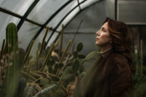 Σε ένα θερμοκήπιο με κάκτους, μια καλοχτενισμένη γυναίκα με καφέ γούνινο πανωφόρι στέκεται προφίλ κοιτώντας ψηλά μπροστά της σαν μια σκέψη να την έχει απορροφήσει και να έχει αφαιρεθεί