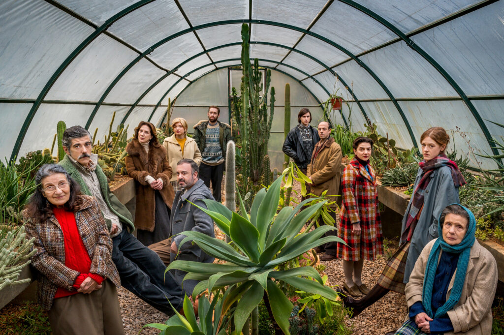 Σε ένα μακρόστενο θερμοκήπιο με κάκτους, παχύφυτα και άλλες πρασινάδες, 11 άτομα απλώνονται κατά μήκος αριστερά και δεξιά από τα φυτά. Φορούν χειμωνιάτικα ρούχα και οι περισσότεροι κοιτούν τον φακό καταβεβλημένοι ή σκυθρωποί.