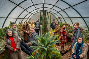 Σε ένα μακρόστενο θερμοκήπιο με κάκτους, παχύφυτα και άλλες πρασινάδες, 11 άτομα απλώνονται κατά μήκος αριστερά και δεξιά από τα φυτά. Φορούν χειμωνιάτικα ρούχα και οι περισσότεροι κοιτούν τον φακό καταβεβλημένοι ή σκυθρωποί.