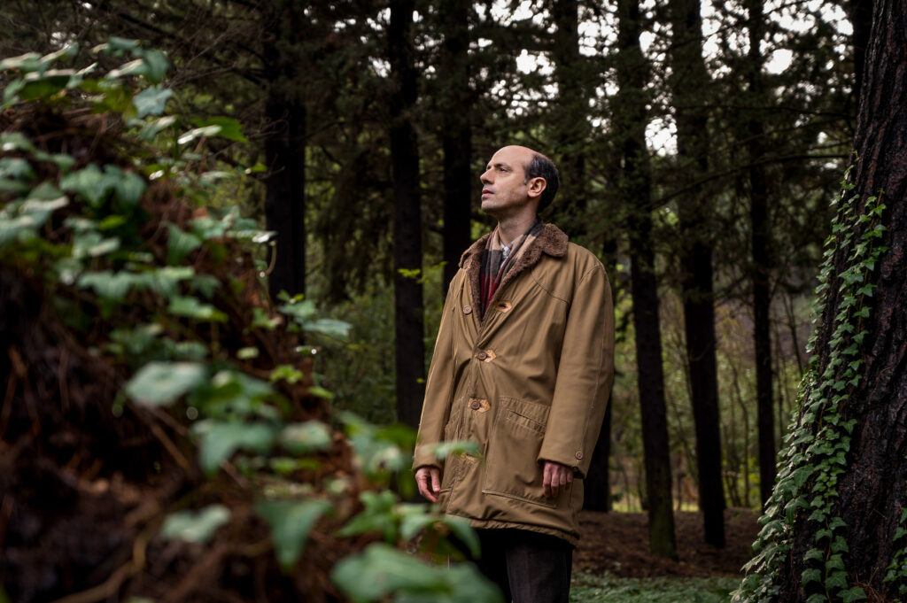 Σε ένα πυκνό δάσος, ένας καραφλός άνδρας με καφέ πανωφόρι ατενίζει στοχαστικά μακριά μπροστά του.