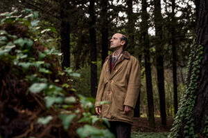 Σε ένα πυκνό δάσος, ένας καραφλός άνδρας με καφέ πανωφόρι ατενίζει στοχαστικά μακριά μπροστά του.