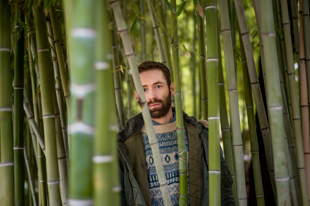 Ανάμεσα σε πυκνούς κορμούς μπαμπού ξεπροβάλλει το πρόσωπο ενός νεαρού άνδρα. Φορά βαρύ πανωφόρι και πλεκτό πουλόβερ και έχει καστανά μαλλιά και μούσια. Με τα μπλε μάτια του κοιτά βαθιά τον φακό αφήνοντας να διαφανεί μια έγνοια ή ανησυχία.