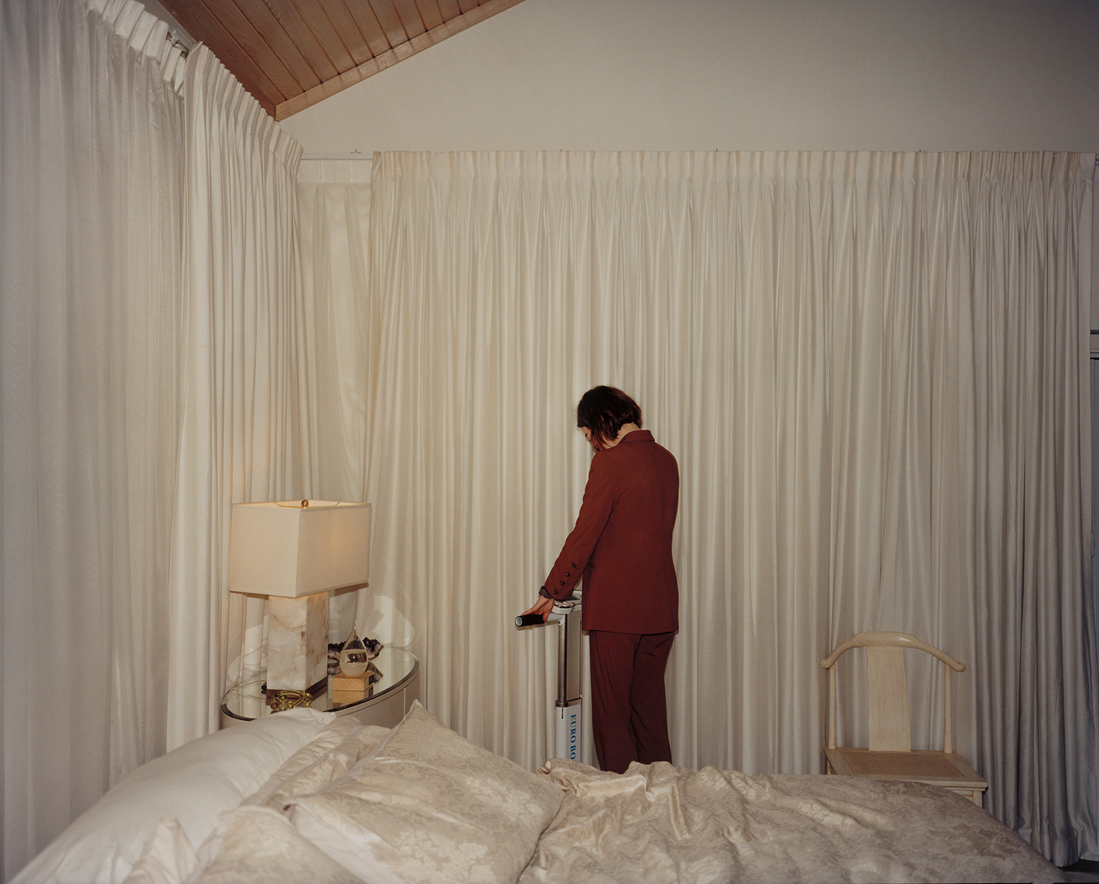 Έγχρωμη φωτογραφία μιας γυναίκας με σκούρο πορτοκαλί κοστούμι σε εσωτερικό χώρο. Η γυναίκα στέκεται πάνω σε ένα μηχάνημα που κρύβεται πίσω από ένα μεγάλο κρεβάτι και θυμίζει πατίνι ή μεγάλη ζυγαριά. Κρατά τις λαβές του και κοιτά προς τα κάτω με πλάτη σε μας. Δίπλα από το κρεβάτι και στο πλάι της γυναίκας υπάρχει ένα κομοδίνο με λευκό πορτατίφ. Οι τοίχοι ολόγυρα είναι καλυμμένοι με λευκή κουρτίνα και το κρεβάτι με λευκά σκεπάσματα, κάνοντας την κοκκινομάλλα γυναίκα με το κεραμιδί της κοστούμι να ξεχωρίζει στο κέντρο της εικόνας.