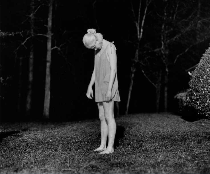Στο σκοτεινό ασπρόμαυρο τοπίο όπου μόνο λεπτοί κορμοί δέντρων ξεχωρίζουν στο βάθος, μια ξανθιά γυναίκα με ένα καλοκαιρινό κοντό φόρεμα στέκεται όρθια σε στάση τριών τετάρτων, ξυπόλυτη πάνω στο ξερό γρασίδι. Το κεφάλι της σκύβει μπροστά σε μια σχεδόν αφύσικη κάμψη του αυχένα προς τα κάτω.