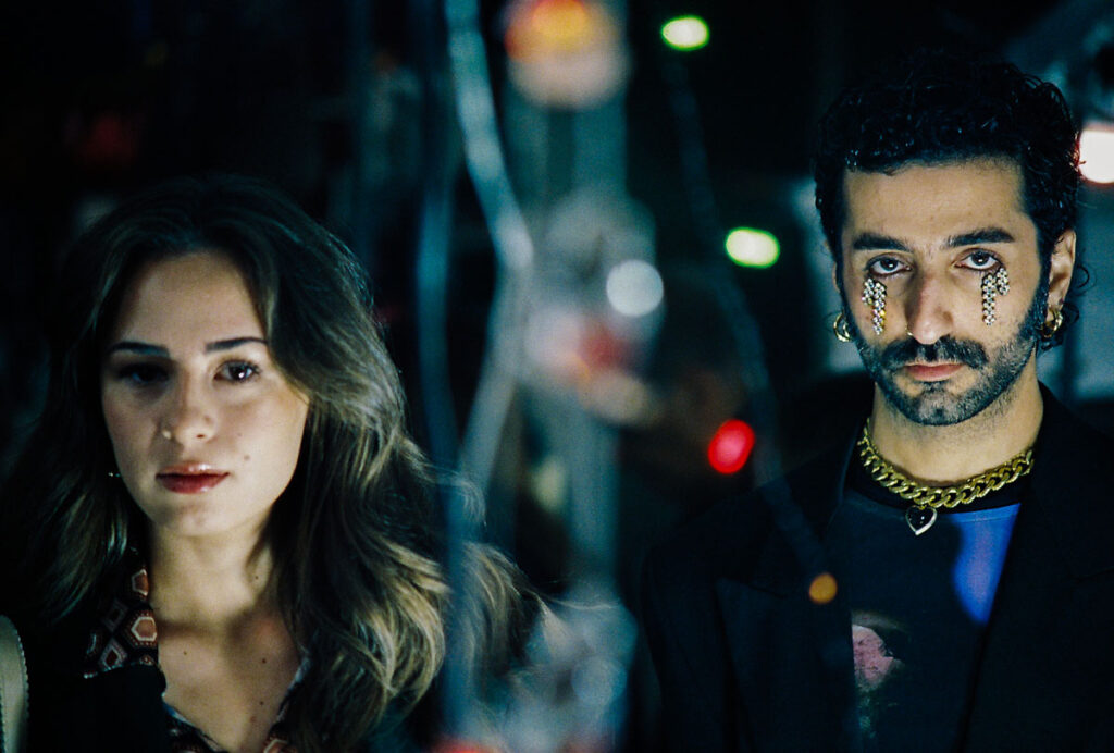 Μια κοπέλα με ανάλαφρα καστανόξανθα μαλλιά και ρόδινα χείλη στέκεται δίπλα από έναν νεαρό μελαχρινό άνδρα με μούσια και χρυσά κοσμήματα. Κάτω από τα μάτια του λαμπυρίζουν πετράδια που παίρνουν τη μορφή δακρύων.