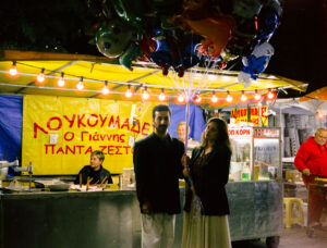 Μπροστά από ένα κίτρινο υπαίθριο σταντ που πουλά λουκουμάδες και ποπ κορν, ένας μελαχρινός άνδρας με μούσια, μαύρο σακάκι και μπεζ παντελόνι και μια νεαρή κοπέλα με αεράτη μπεζ μακριά φούστα και μαύρο σακάκι κρατούν ένα μάτσο μπαλόνια με ήλιον που ανεβαίνουν προς τα πάνω και χάνονται στο όριο της φωτογραφίας.