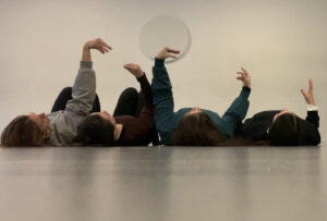 4 γυναίκες είναι ξαπλωμένες η μια δίπλα στην άλλη στο γκρίζο πάτωμα ενός φωτεινού χώρου με λευκό τοίχο. Βλέπουμε τις κορυφές των κεφαλιών τους και τα χέρια τους υψωμένα να σχηματίζουν με τα δάχτυλα και τον αντίχειρα το ράμφος ίσως κάποιου πτηνού.