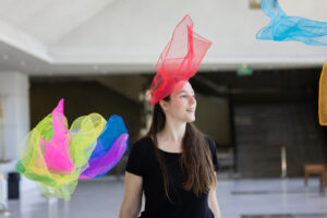 Σε απλόχωρο εσωτερικό χώρο, μια νεαρή γυναίκα με μαύρο καλοκαιρινό μπλουζάκι και καστανόξανθα μακριά μαλλιά κινείται ανάμεσα σε αιωρούμενα αραχνοϋφαντα μαντήλια σε πολλά ζωντανά χρώματα. Το πρόσωπό της γαλήνιο ατενίζει πλάγια μπροστά της την κίνηση των μαντηλιών στον χώρο γύρω της.