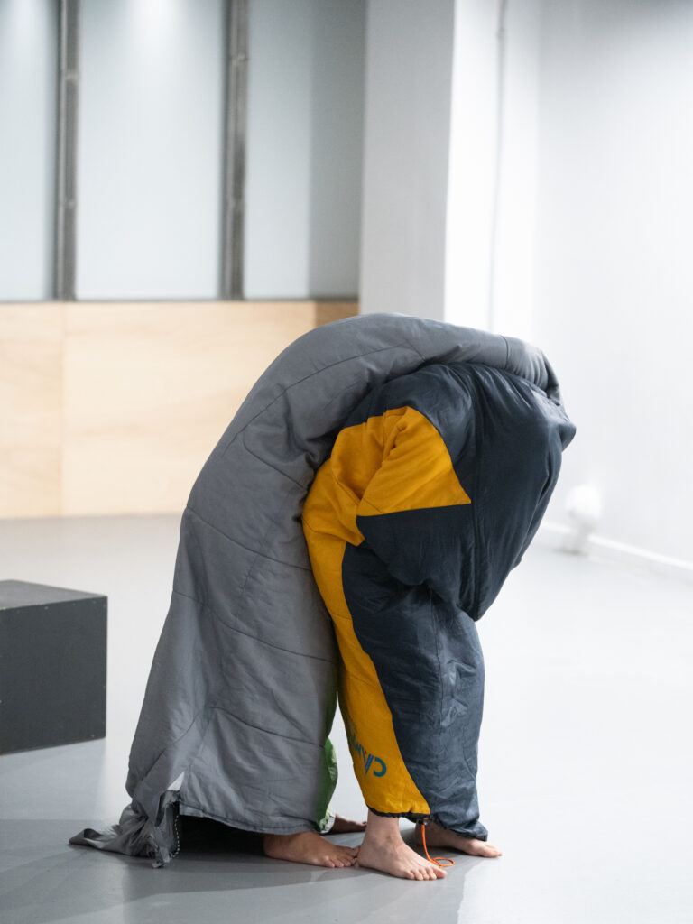 Σε έναν φωτεινό χώρο με γκρι ανοιχτό γυαλιστερό πάτωμα, δύο σώματα είναι κρυμμένα κάτω από σκουρόχρωμους υπνόσακους. Στέκονται το ένα πίσω από το άλλο. Καθώς το πίσω σώμα έχει γείρει μπροστά πάνω στο άλλο, το μπροστινό σκύβει μπροστά για να το στηρίξει. Πίσω τους ένας τετράγωνος μαύρος κύβος είναι το μοναδικό αντικείμενο στον χώρο.