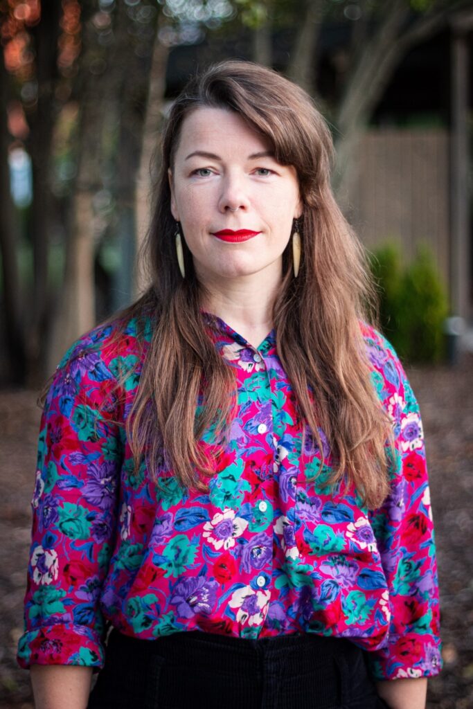 Έγχρωμη φωτογραφία πορτρέτου μιας γυναίκας που στέκεται σε υπαίθριο χώρο. Έχει μακριά καστανά μαλλιά με πλαϊνές αφέλειες, κόκκινο κραγιόν και σκουλαρίκι στη μύτη. Φορά μεγάλα, ανοιχτόχρωμα κρεμαστά σκουλαρίκια και ένα πολύχρωμο φλοράλ μπλουζάκι (ροζ, μωβ και τιρκουάζ), συνδυασμένο με μαύρο ψηλόμεσο παντελόνι.