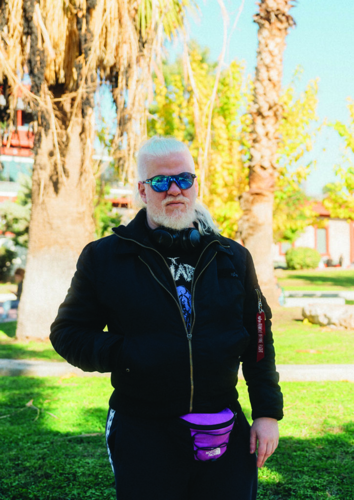 Έγχρωμη φωτογραφία πορτρέτου ενός νεαρού κατάξανθου άνδρα σε υπαίθριο ηλιόλουστο χώρο που θυμίζει πάρκο. Ο άνδρας έχει μακριά μαλλιά πιασμένα σε ψηλή κοτσίδα και πυκνά γένια. Τα μάτια του κρύβονται πίσω από στρογγυλά γυαλιά ηλίου με καθρέφτη. Ακουστικά τυλίγουν τον λαιμό του και ένα φούξια τσαντάκι μέσης ξεχωρίζει πάνω στα μαύρα του ρούχα. Φορά μπουφάν με φερμουάρ και φόρμα και έχει το ένα του χέρι στην τσέπη καθώς κοιτά με σοβαρή έκφραση προς τον φακό.